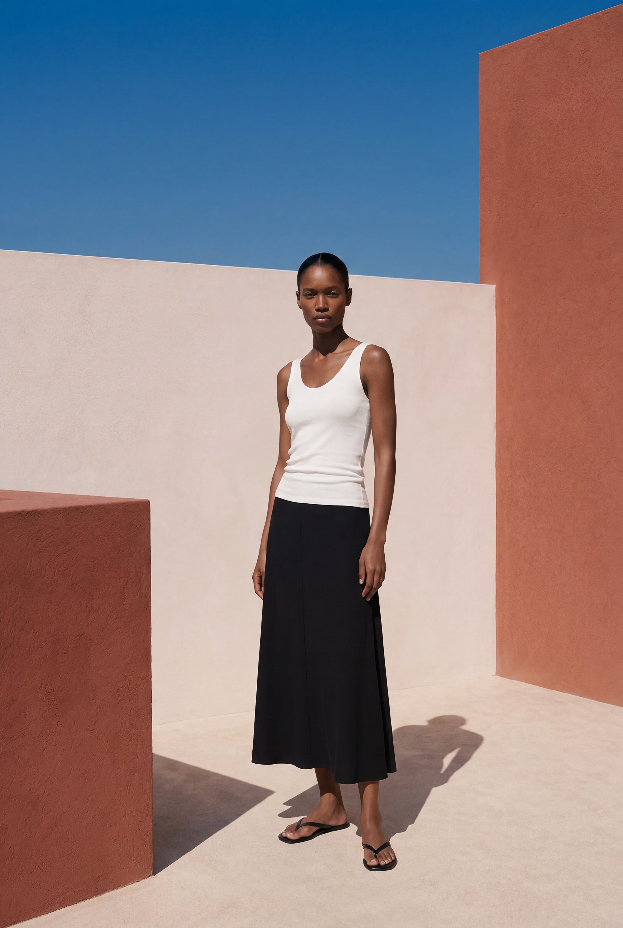 Woman in a white tank top and black skirt standing against a geometric wall with blue sky.